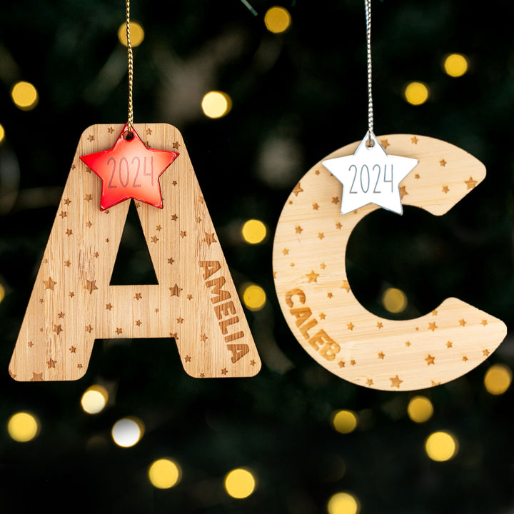 Two bamboo Christmas Twinkle Letter Baubles, shaped as A and C, hang on a tree with engraved names (AMELIA, CALEB), 2024 star tags, and small stars. Blurred festive lights glow behind these personalised ornaments.