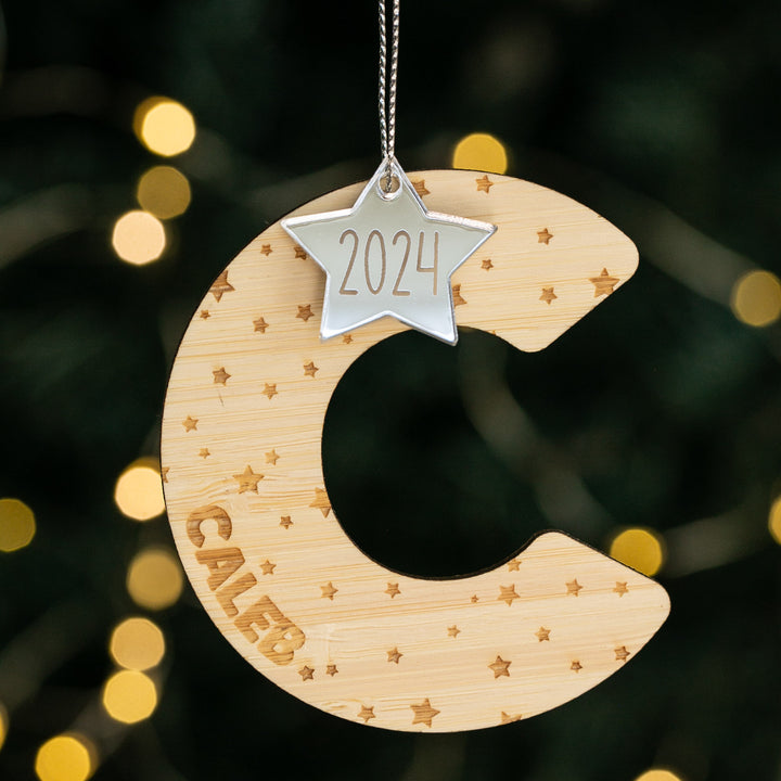 A natural bamboo Christmas ornament with a personalized engraving of a name and the year 2024, adorned with a silver star and small dangling stars.
