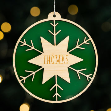 The Winter Star Christmas Bauble is a round wooden ornament with a green background, laser-cut snowflake design, and "Thomas" engraved at the center, accented by blurred festive lights in the holiday décor.