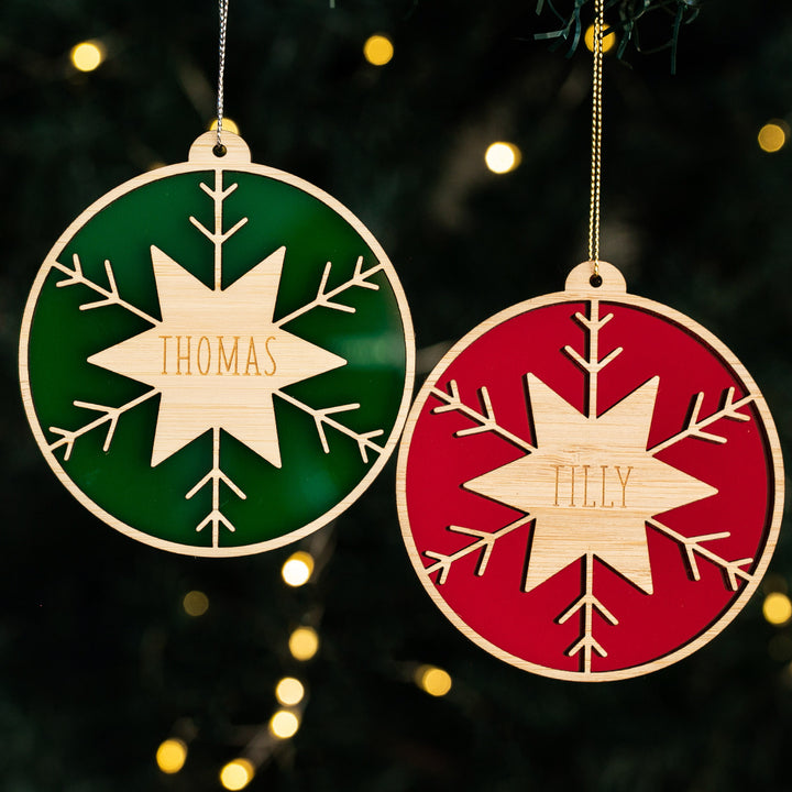 Two Winter Star Christmas Baubles, one green engraved with THOMAS and one red with TILLY, feature snowflake designs and hang against a dark background with fairy lights—perfect holiday décor.