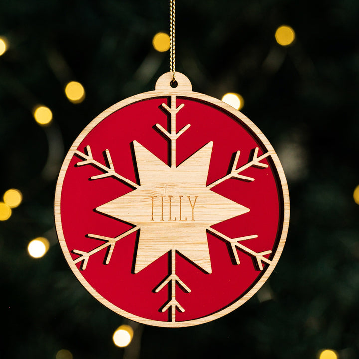 The Winter Star Christmas Bauble is a personalised wooden ornament with a red background, snowflake design, and laser-cut center star engraved with "TILLY"—a charming addition to your holiday décor.