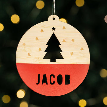 The Woodland Christmas Bauble is a wooden ornament with a tree and stars above “JACOB,” festive red bottom, and hangs beautifully in front of warm holiday lights—ideal for personalised Christmas decorating.