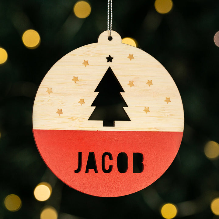 The Woodland Christmas Bauble is a wooden ornament with a tree and stars above “JACOB,” festive red bottom, and hangs beautifully in front of warm holiday lights—ideal for personalised Christmas decorating.