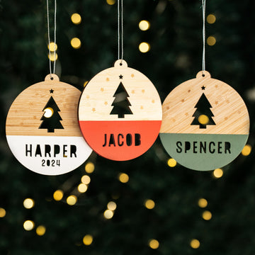 Three Woodland Christmas Baubles hang side by side—featuring a cutout tree, stars, and personalised names: Harper 2024 on white, Jacob on red, and Spencer on green—set against softly blurred holiday lights.
