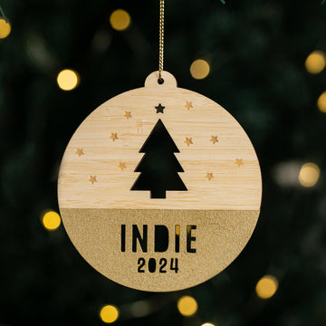 The Woodland Christmas Bauble features a cut-out tree and stars, gold glitter base, and “INDIE 2024” text, hanging in front of blurred Christmas lights—a delightful personalised festive decoration.