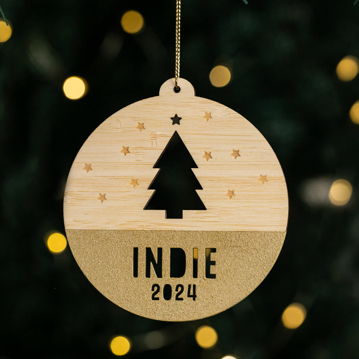 The Woodland Christmas Bauble features a cut-out tree and stars, gold glitter base, and “INDIE 2024” text, hanging in front of blurred Christmas lights—a delightful personalised festive decoration.