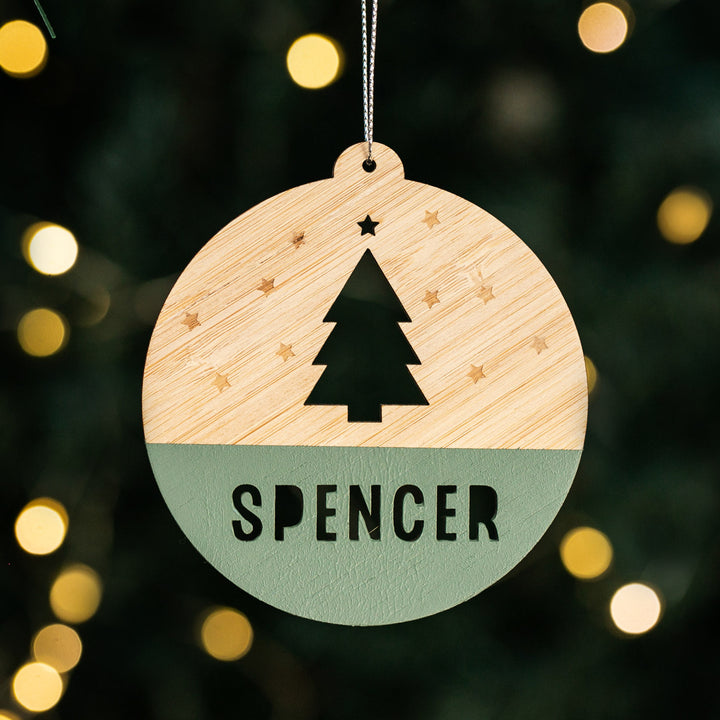 Our Woodland Christmas Bauble is a round bamboo ornament featuring a cut-out tree, star accents, and SPENCER at the bottom. It hangs beautifully on your tree, catching festive lights for a magical touch.