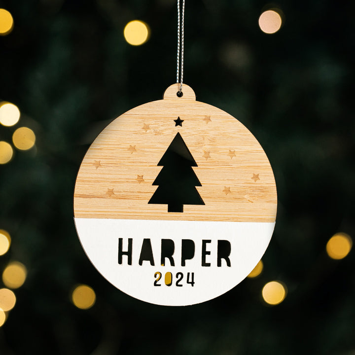 A round wooden Woodland Christmas Bauble with a cutout tree, star accents, and "Harper 2024" engraved, hanging against blurred holiday lights.