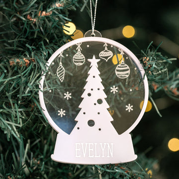 The Personalised Christmas Snowglobe ornament features a cutout Christmas tree with ornaments and snowflakes, plus the name EVELYN at the base—a festive and unique decoration for your holiday tree.