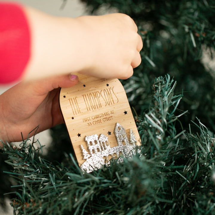 A child hangs the "Our First Home Christmas Globe," a personalised bauble with engraved text and silver glitter, on a green tree. It reads "THE THOMPSONS FIRST CHRISTMAS AT 23 CLYDE STREET"—a perfect new home ornament.