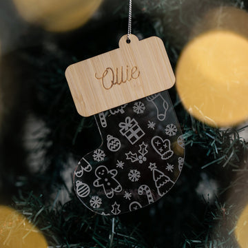 A Christmas Stocking Bauble, clear with white holiday designs and a wooden top labeled Ollie, hangs on a tree among green needles and warm lights—ideal for fans of personalised Christmas ornaments.