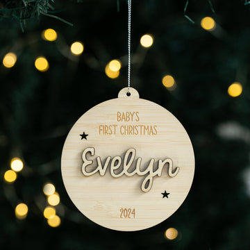 The Celebration Christmas Bauble is a round, personalised ornament made from eco-friendly bamboo, engraved with BABYS FIRST CHRISTMAS Evelyn 2024 and star accents, hanging on a tree with blurred yellow holiday lights in the background.