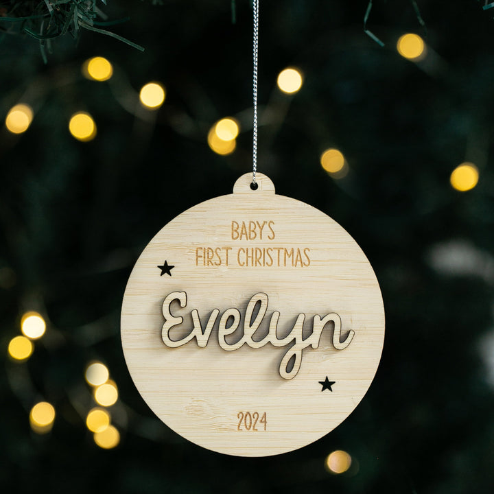 The Celebration Christmas Bauble is a round, personalised ornament made from eco-friendly bamboo, engraved with BABYS FIRST CHRISTMAS Evelyn 2024 and star accents, hanging on a tree with blurred yellow holiday lights in the background.