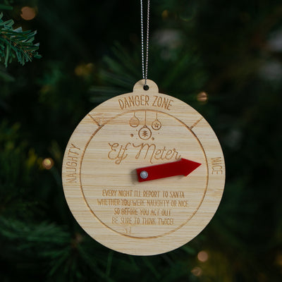 A round wooden Elf Meter Christmas ornament hanging from a silver string against a blurred Christmas tree background. The natural wood disc features engraved text and a red pointer arrow.