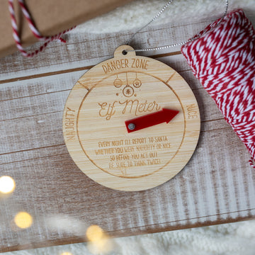 A round wooden Elf Meter ornament with engraved text and a red arrow pointer, styled on a rustic wooden surface with red-and-white bakers twine and gift packaging.