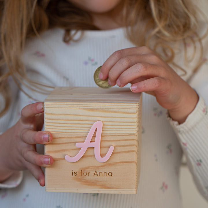 A young child wearing a white floral shirt puts a coin into the Personalised Wooden Money Box, which features a large pink letter A and the words "is for Anna" on it.