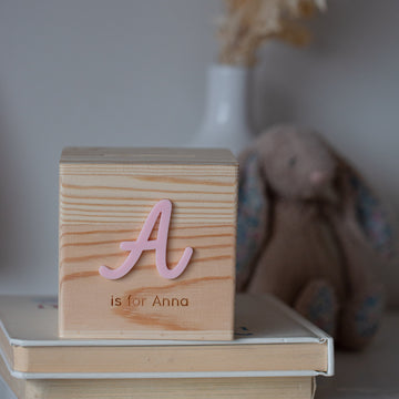 A wooden block with a pink letter A and "is for Anna" sits atop stacked books, next to a Personalised Wooden Money Box—an ideal keepsake gift—with a plush bunny and vase softly blurred in the background.