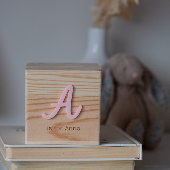 A wooden block with a pink letter A and "is for Anna" sits atop stacked books, next to a Personalised Wooden Money Box—an ideal keepsake gift—with a plush bunny and vase softly blurred in the background.