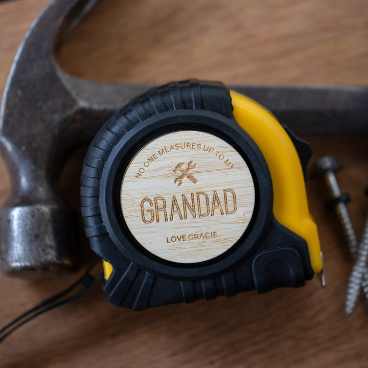 A yellow and black Personalised Tape Measure with a wooden label saying "No one measures up to my Grandad. Love Gracie." sits by a hammer and screws—a unique Father’s Day gift and custom toolbox essential—on a wooden surface.