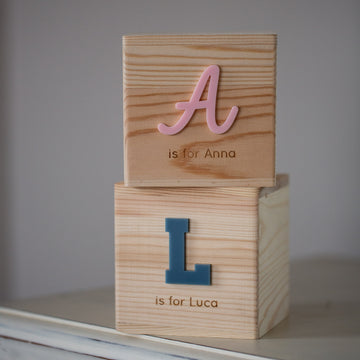 Two Personalised Wooden Money Boxes are stacked: the top has a pink "A is for Anna," the bottom a blue "L is for Luca." Displayed on a light surface, they make a charming keepsake gift.