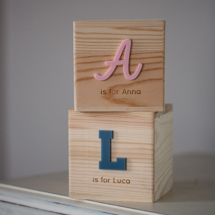 Two Personalised Wooden Money Boxes are stacked: the top has a pink "A is for Anna," the bottom a blue "L is for Luca." Displayed on a light surface, they make a charming keepsake gift.