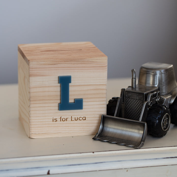 A Personalised Wooden Money Box featuring a blue letter L and "is for Luca" rests on a light surface beside a small metal toy bulldozer.