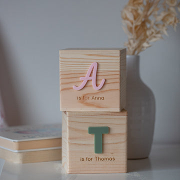 A Personalised Wooden Money Box with pink "A" and green "T" blocks is stacked for Anna and Thomas. A white vase with dried flowers in the background completes this charming keepsake gift.