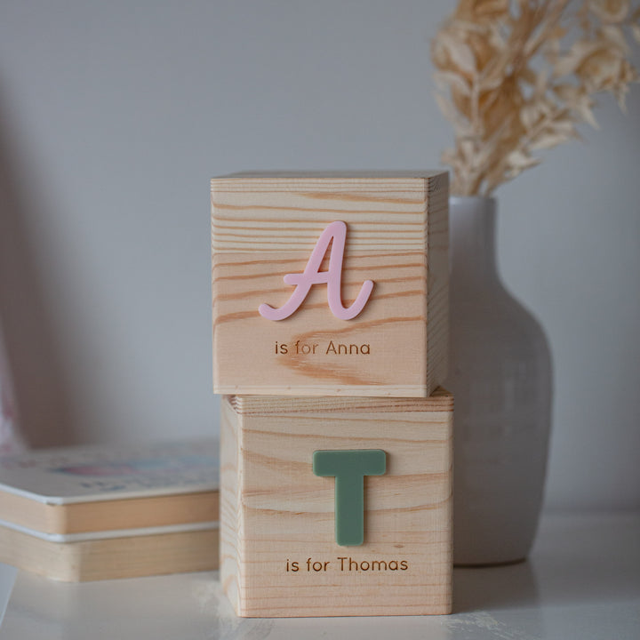 A Personalised Wooden Money Box with pink "A" and green "T" blocks is stacked for Anna and Thomas. A white vase with dried flowers in the background completes this charming keepsake gift.