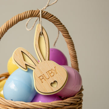 A wicker basket with pastel eggs features a Personalised Bunny Tales Easter Tag, engraved with the name RUBY and a small heart cutout on the handle—an ideal addition to your Easter collection.