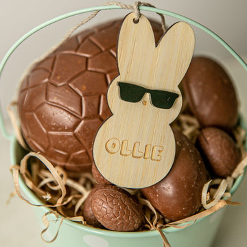 A mint-green bucket comes with large and small chocolate eggs, straw, and a bamboo Personalised Happy Peeps Easter Tag engraved with OLLIE. The bunny tag wears black sunglasses for a playful Easter touch to your collection.