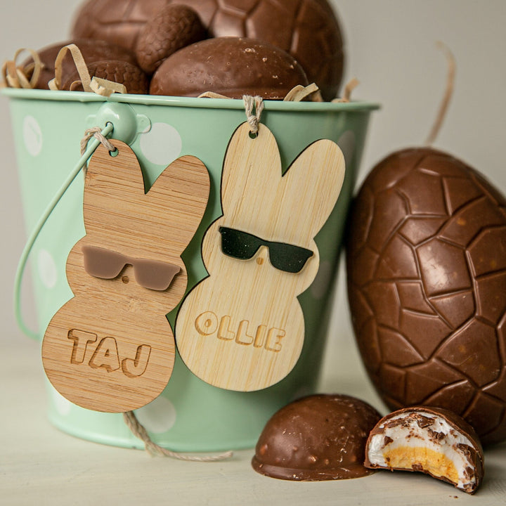 A green polka dot bucket filled with chocolate eggs and candies features eco-friendly Personalised Happy Peeps Easter Tags labeled TAJ and OLLIE with sunglass-wearing bunnies. A broken, creamy-filled chocolate egg sits in front for extra charm.