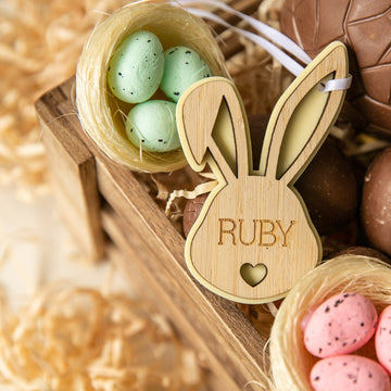 A Personalised Bunny Tales Easter Tag engraved with RUBY hangs on a crate next to pastel green and pink speckled eggs in small nests, surrounded by shredded paper and chocolate eggs—a charming Easter collection addition.