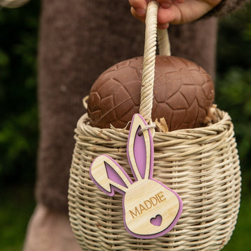 A hand holds a wicker basket with a large chocolate egg. Attached is a Personalised Bunny Tales Easter Tag shaped like a bunny, featuring the name Maddie in pink. Greenery from our Easter collection appears in the background.