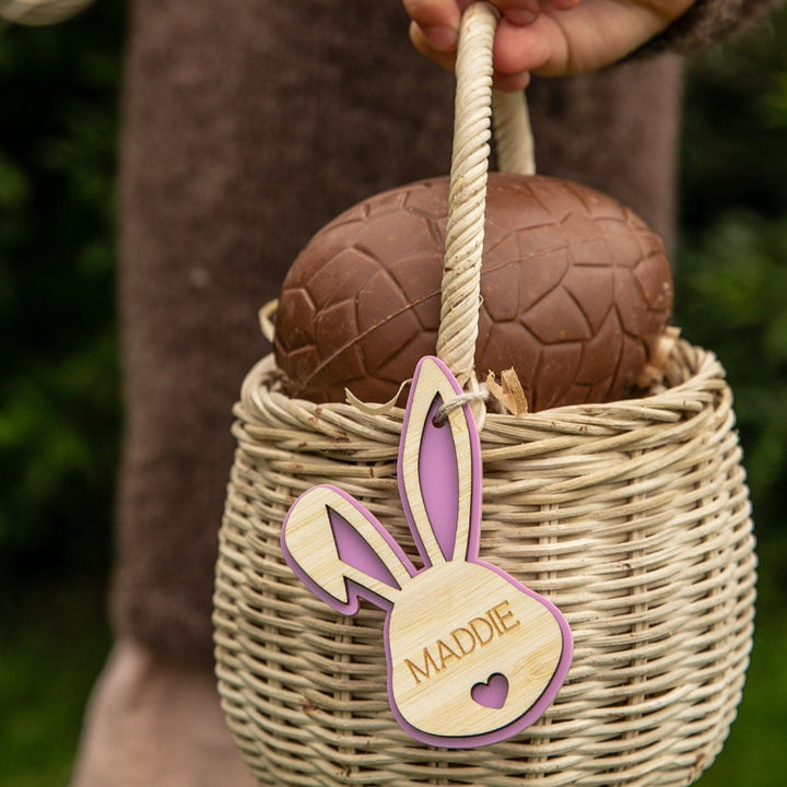 A hand holds a wicker basket with a large chocolate egg. Attached is a Personalised Bunny Tales Easter Tag shaped like a bunny, featuring the name Maddie in pink. Greenery from our Easter collection appears in the background.