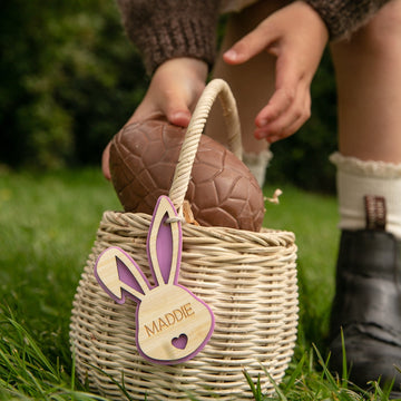 A child puts a large chocolate egg into a basket decorated with a Personalised Bunny Tales Easter Tag labeled MADDIE, while wearing socks and black shoes. Grass and greenery can be seen in the background.