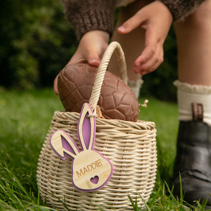 A child puts a large chocolate egg into a basket decorated with a Personalised Bunny Tales Easter Tag labeled MADDIE, while wearing socks and black shoes. Grass and greenery can be seen in the background.