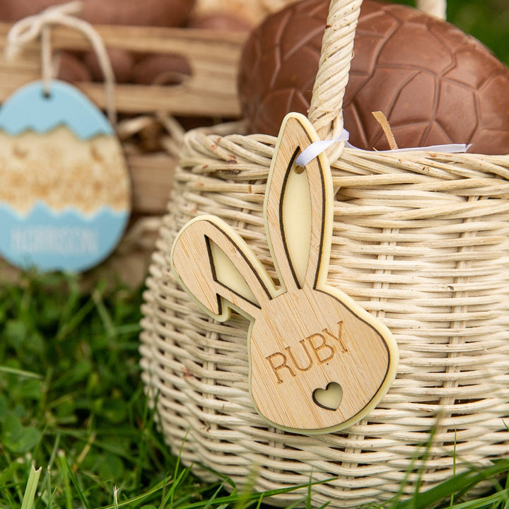 A close-up of a wicker basket with a Personalised Bunny Tales Easter Tag, engraved with RUBY and a heart, holding a chocolate egg on green grass. Another basket with a blue tag appears blurred in the background.