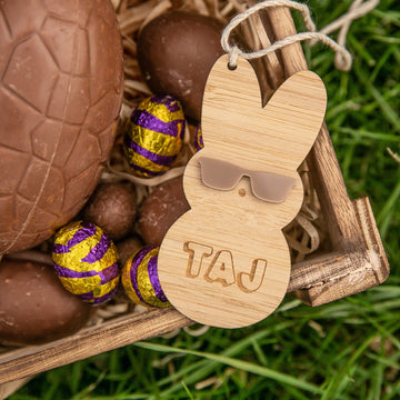 The Personalised Happy Peeps Easter Tag features an eco-friendly bamboo bunny with sunglasses and TAJ engraving, presented in a wooden box with chocolate eggs—some in purple and gold foil—on straw and grass.