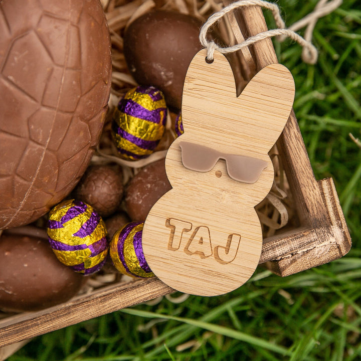 The Personalised Happy Peeps Easter Tag features an eco-friendly bamboo bunny with sunglasses and TAJ engraving, presented in a wooden box with chocolate eggs—some in purple and gold foil—on straw and grass.