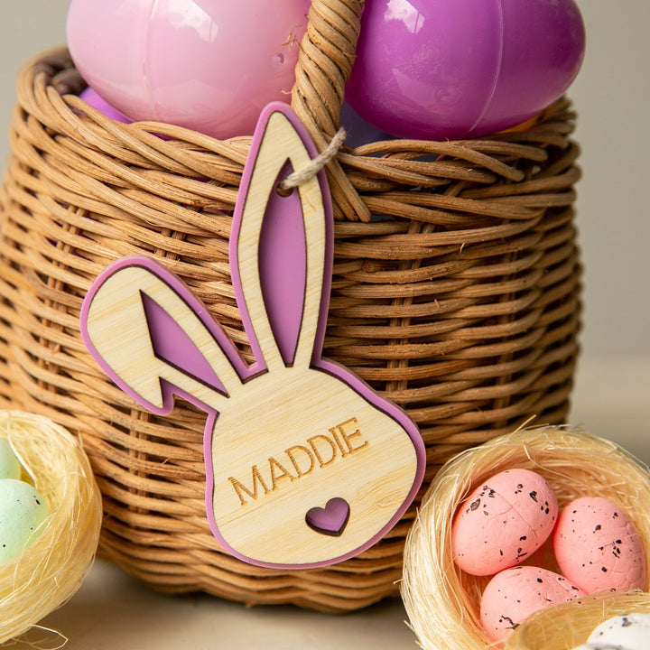 A woven basket holds pastel eggs and features a Personalised Bunny Tales Easter Tag with pink trim and the name MADDIE. Two small nests with colored speckled eggs complete this charming Easter collection.