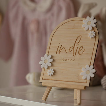The Personalised Daisy Arch Signage, engraved with “indie GRACE” and decorated with white flower accents, stands on a small easel. In the soft background, a blush pink baby outfit hangs—ideal for nursery decor.