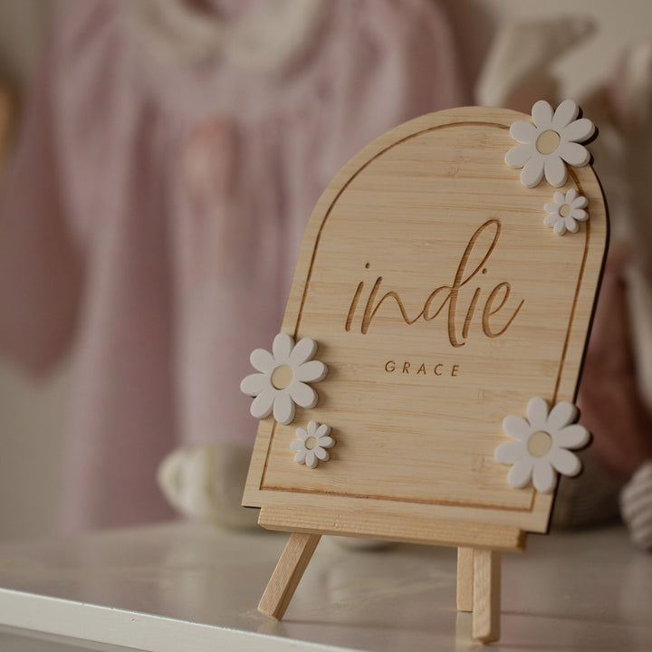 The Personalised Daisy Arch Signage, engraved with “indie GRACE” and decorated with white flower accents, stands on a small easel. In the soft background, a blush pink baby outfit hangs—ideal for nursery decor.