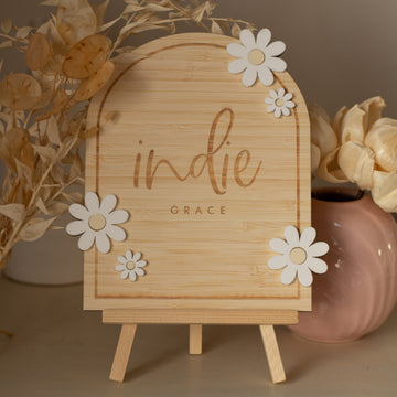 A Personalised Daisy Arch Signage stands on a small easel, decorated with white flower cutouts and surrounded by dried plants and a pink vase holding cream-colored flowers.