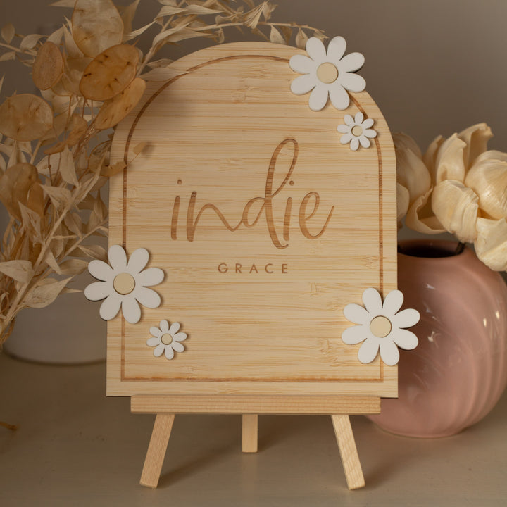 A Personalised Daisy Arch Signage stands on a small easel, decorated with white flower cutouts and surrounded by dried plants and a pink vase holding cream-colored flowers.
