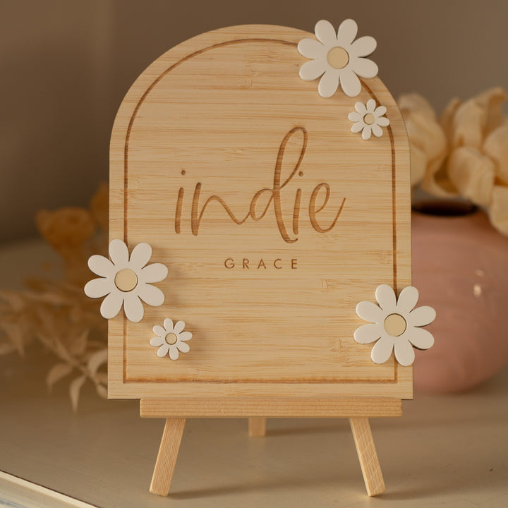 A Personalised Daisy Arch Signage sits on a wooden easel, featuring "indie GRACE" in cursive and block letters. White daisies adorn the custom plaque, with beige flowers and a pink vase softly blurred in the background.