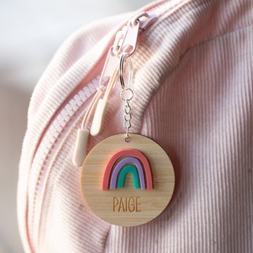 A Rainbow Personalised Bag Tag engraved with Paige hangs from a pink corduroy backpack by the zipper, adding a cheerful touch to any child&
