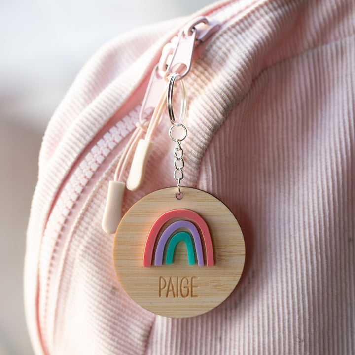A Rainbow Personalised Bag Tag engraved with Paige hangs from a pink corduroy backpack by the zipper, adding a cheerful touch to any child&