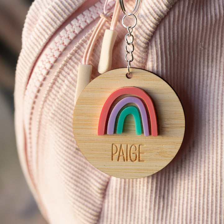A round wooden Rainbow Personalised Bag Tag featuring a colorful rainbow and the name PAIGE engraved, attached to the zipper of a light pink corduroy bag.