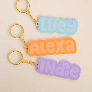 Three Bubblegum Personalised Bag Tags with gold rings and pastel name tags—LUCY in blue, ALEXA in orange, and INDIE in purple—arranged vertically on a white background. Perfect as custom acrylic keyrings.