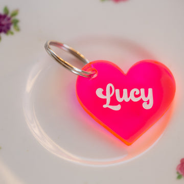 A Personalised Pet Tag, shaped like a bright pink heart with "Lucy" in white lettering, is displayed on a white plate decorated with small floral designs.